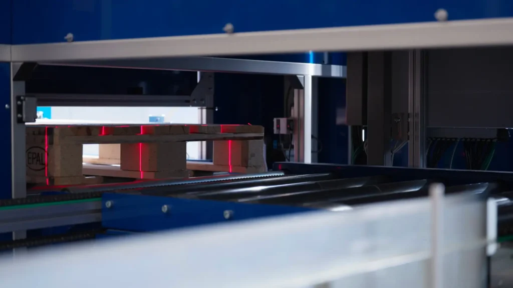 An empty pallet enters an automated pallet inspection station, where a red laser beam scans it for contactless 2D and 3D quality inspection.