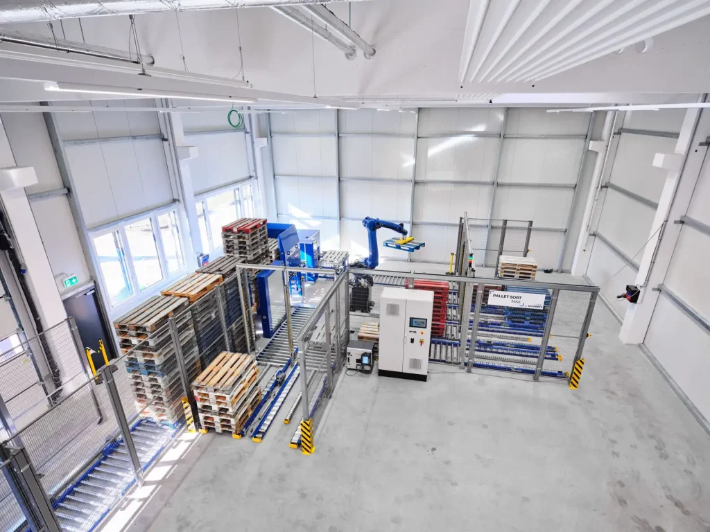 Übersicht über die PalletSort Max Palettensortieranlage von AUTFORCE. Sichtbar sind die Aufabebahn, das Vereinzeln der Paletten, PACO (die Palettenkontrolle mittels 2D und 3D Sensoren und Kameras), der Roboter, der die Paletten auf den richtigen Ausgabebahnen stapelt.